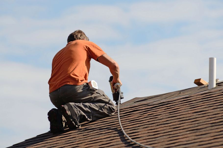 A person wearing an orange shirt kneels on a residential roof, using a pneumatic nail gun to install roofing shingles.