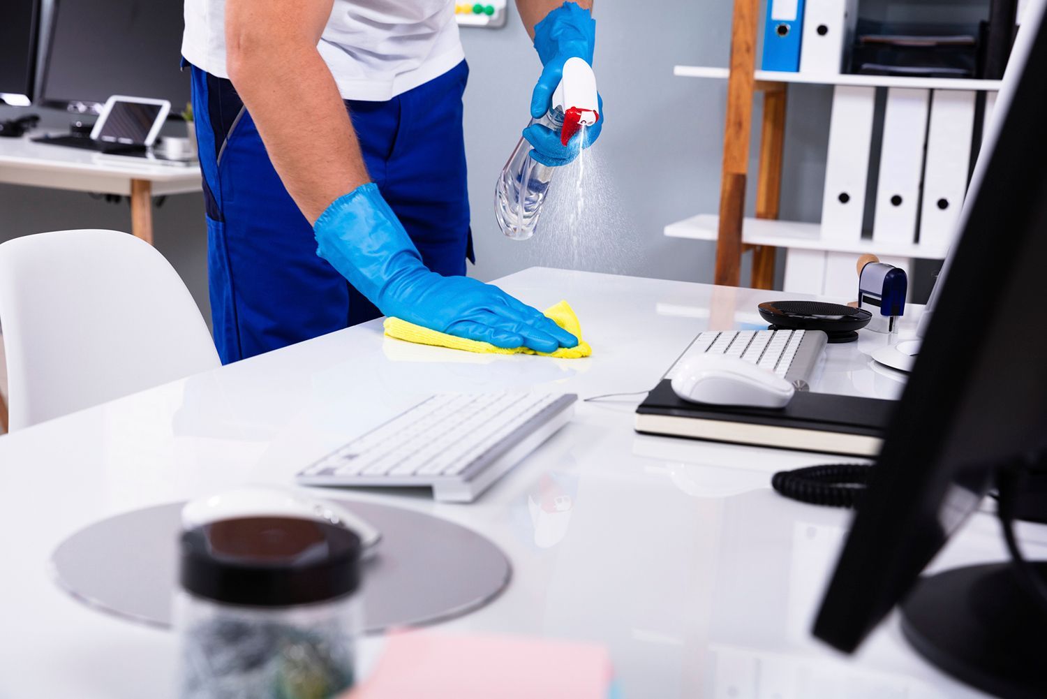 Person wearing blue gloves, cleaning a white office desk with spray and a yellow cloth.