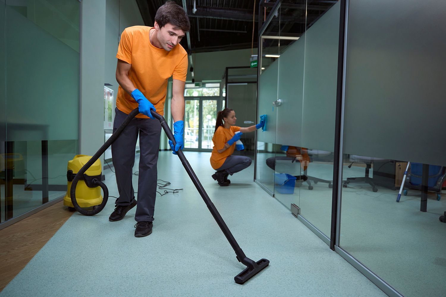 Two people cleaning an office: one vacuuming the floor, the other wiping glass, both wearing gloves and orange shirts. Two people cleaning an office: one vacuuming the floor, the other wiping glass, both wearing gloves and orange shirts.