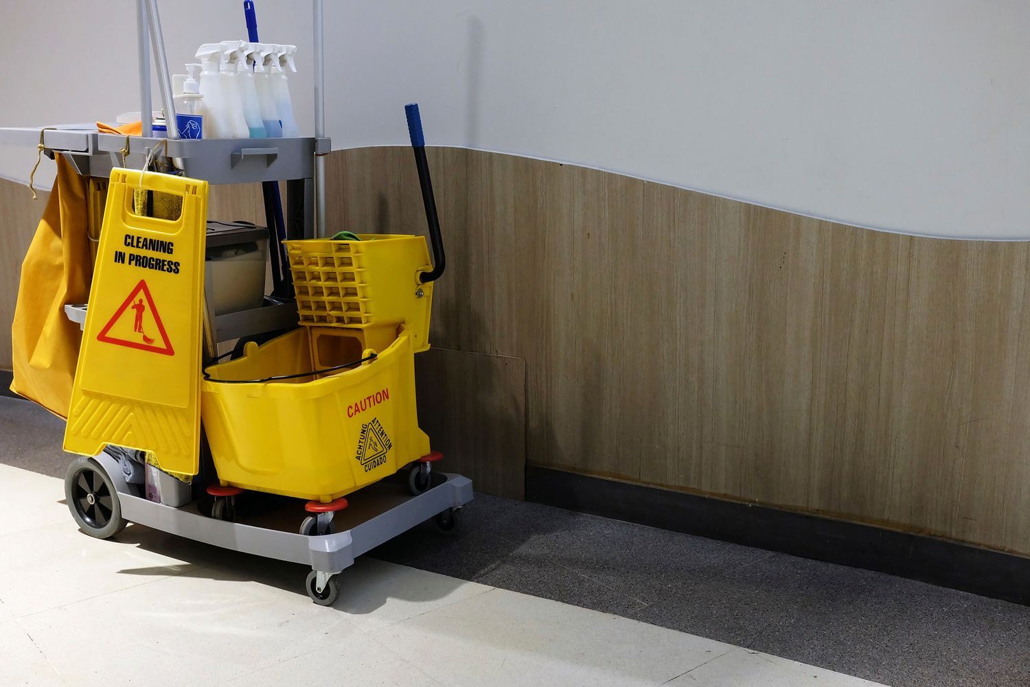 Yellow cleaning cart with warning sign and supplies against a light wood-paneled wall. Yellow cleaning cart with warning sign and supplies against a light wood-paneled wall.