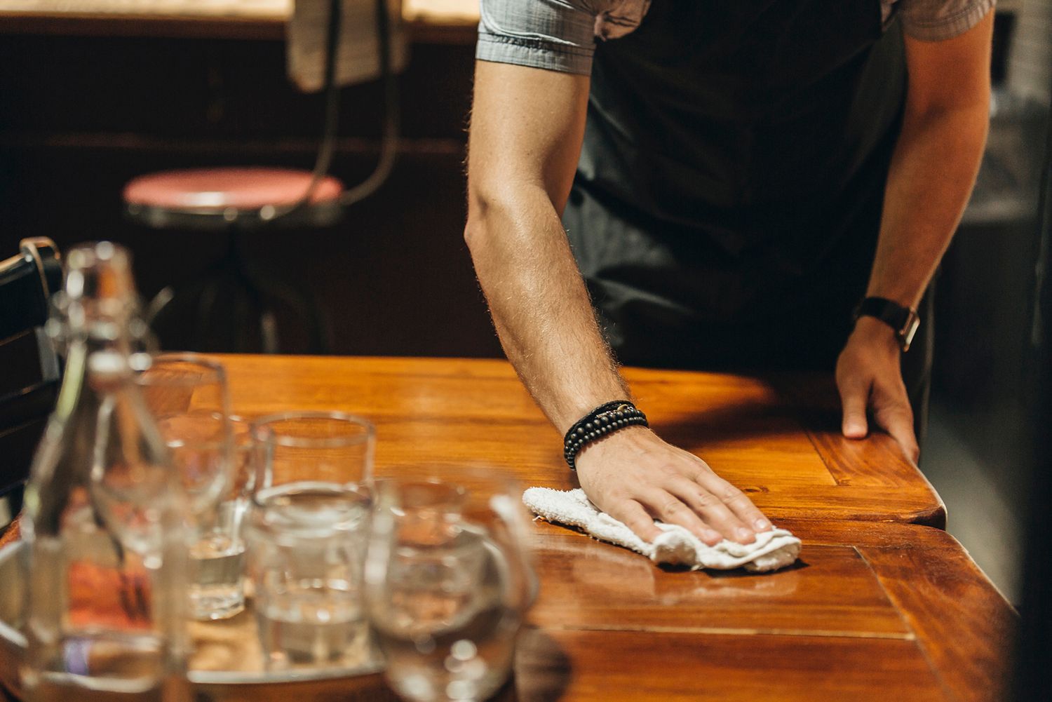 Person wiping a wooden table in a bar; glasses and a stool are visible.