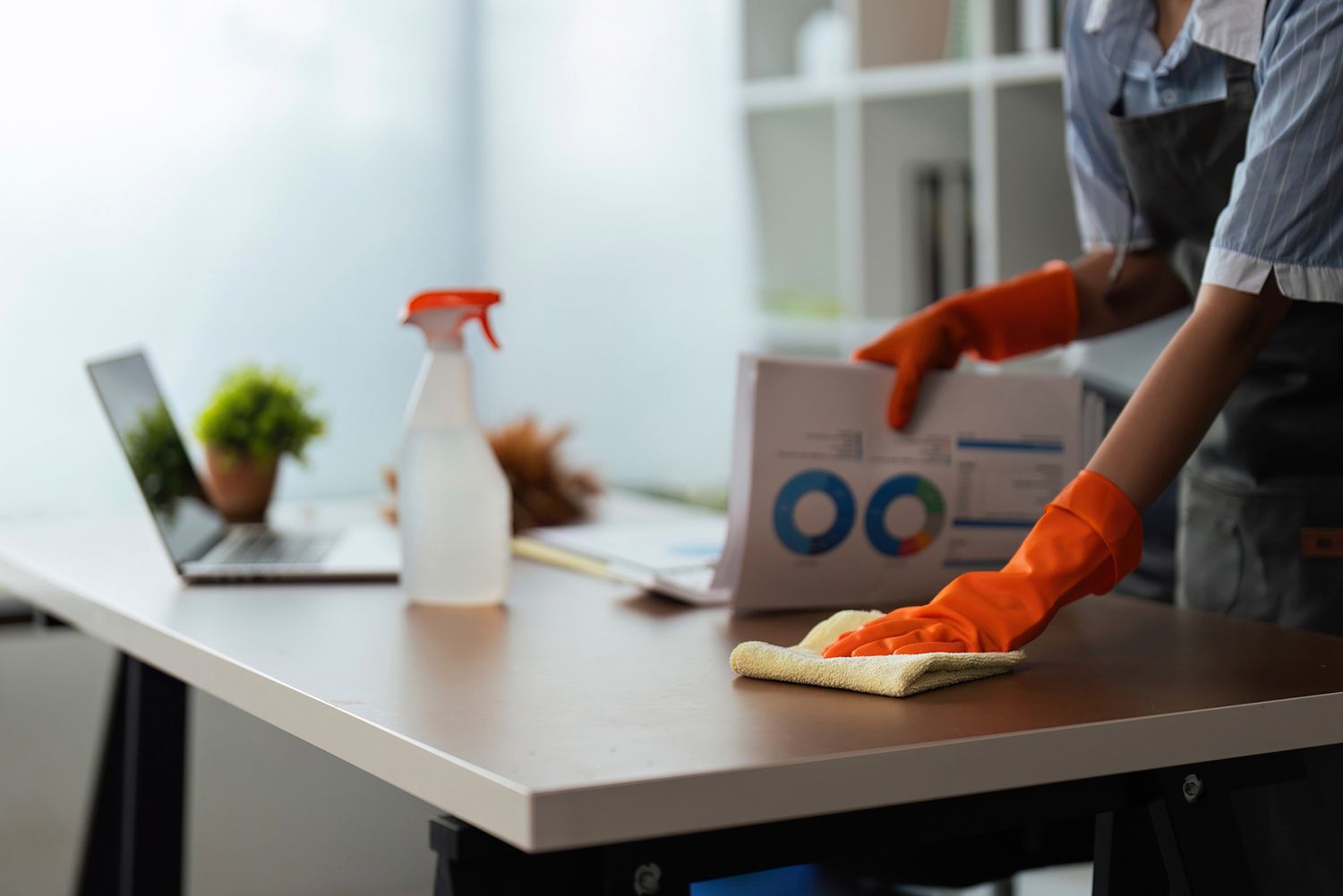Person in orange gloves cleaning a desk in an office. A laptop, spray bottle, and papers are on the desk.