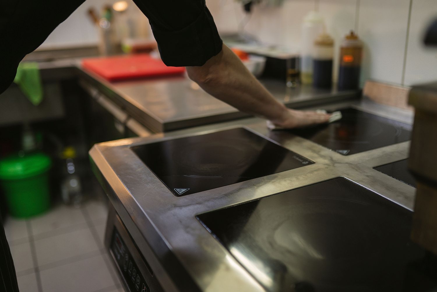 Person wiping a black stovetop in a commercial kitchen with stainless steel counters.