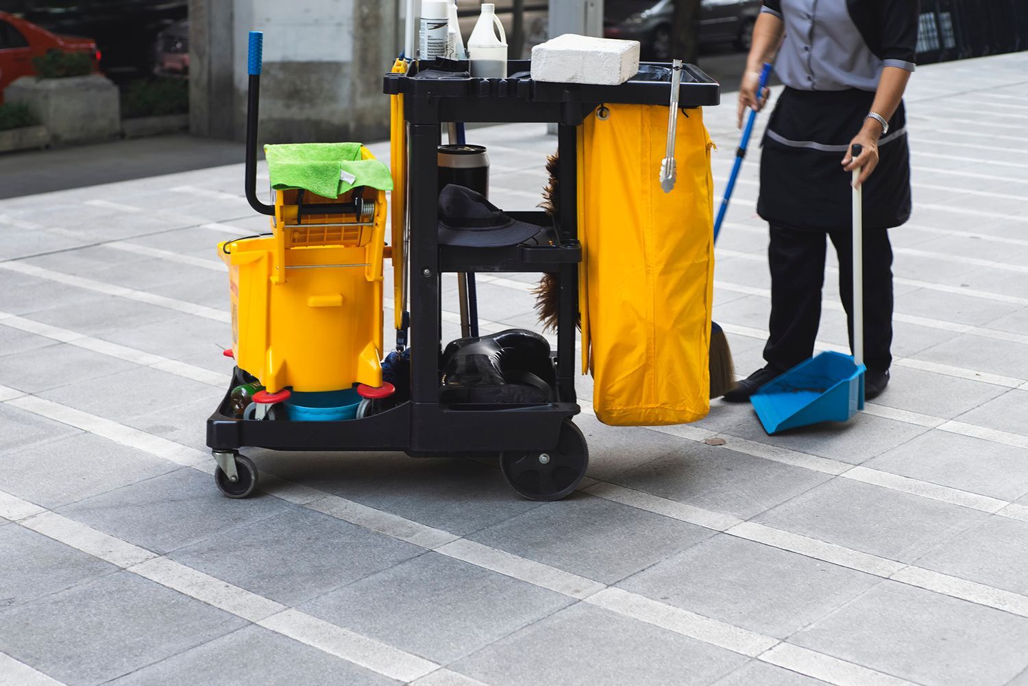 A person sweeping an outdoor walkway; a cleaning cart is nearby.