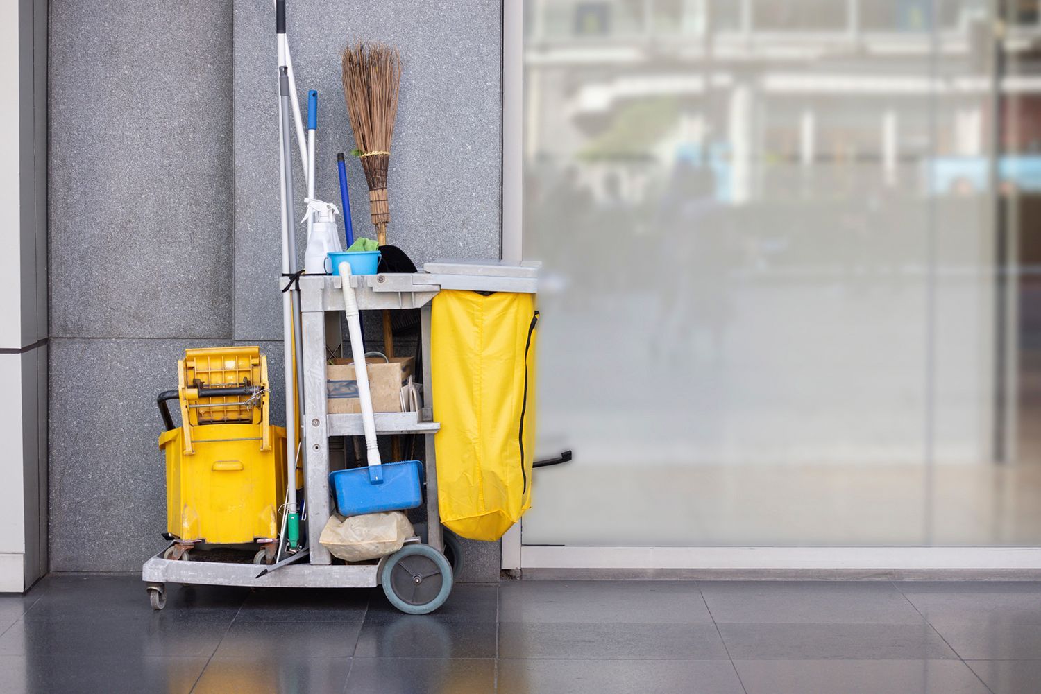 Cleaning supplies cart next to a building. Yellow bucket, broom, mop, blue mop, and yellow trash bag. Cleaning supplies cart next to a building. Yellow bucket, broom, mop, blue mop, and yellow trash bag.