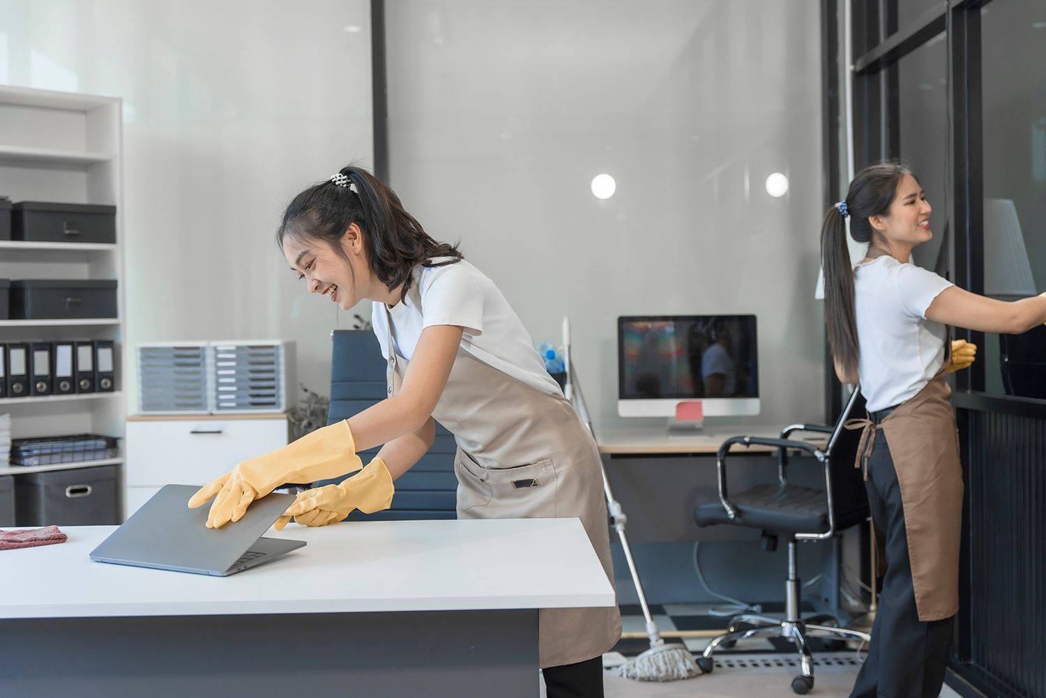 Two people cleaning an office. One wipes a laptop, the other cleans a window. Both wear aprons and gloves.