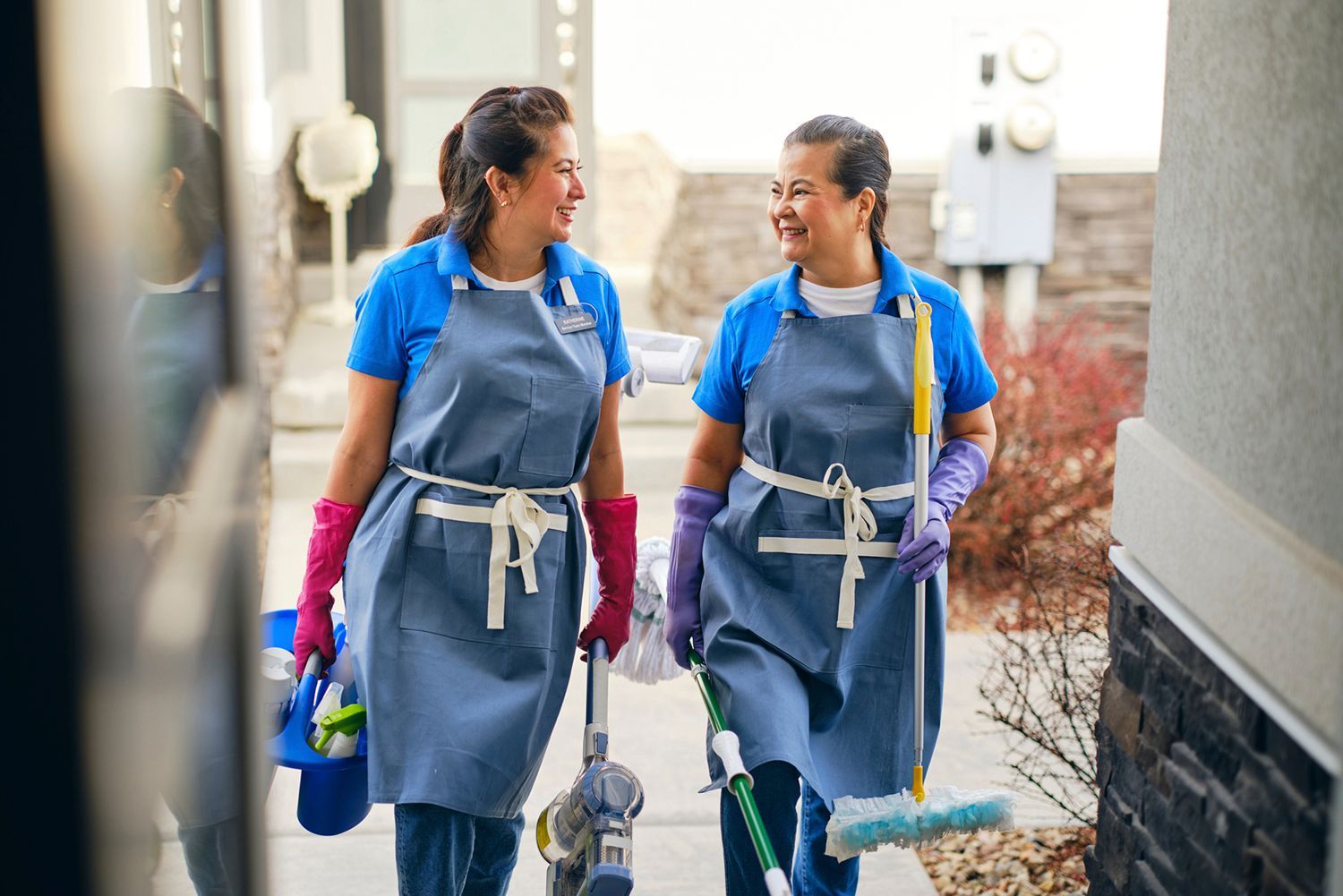 Two cleaning service workers walk together smiling, carrying cleaning supplies.