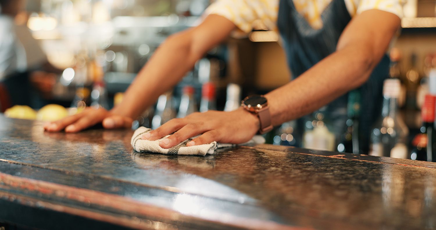 Man vacuuming floor in a cafe, lifting a wooden chair, wearing apron and shorts.