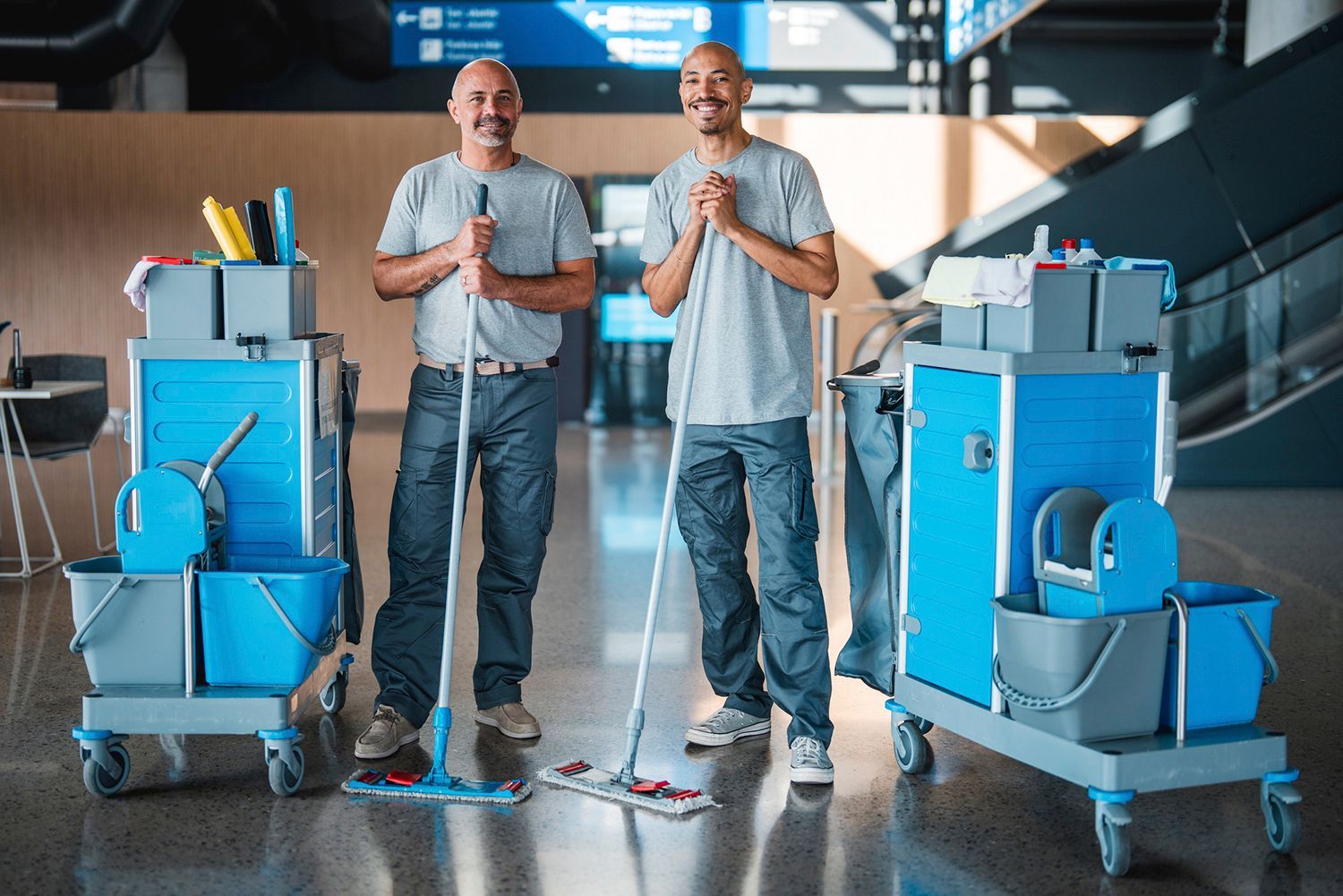 Two men, with cleaning carts, stand indoors. Smiling, they pose with mops in front of an airport interior. Two men, with cleaning carts, stand indoors. Smiling, they pose with mops in front of an airport interior.