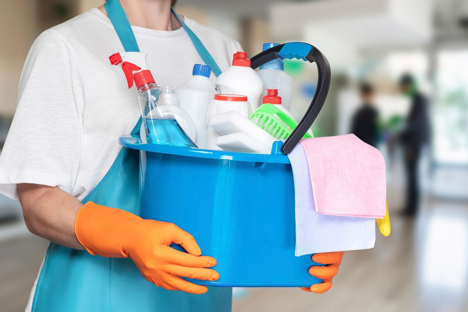Person wearing apron and orange gloves, holding a blue bucket filled with cleaning supplies. Person wearing apron and orange gloves, holding a blue bucket filled with cleaning supplies.