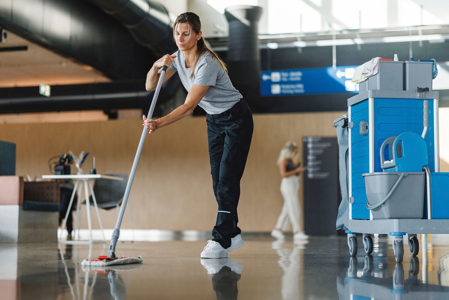 Woman mopping a shiny floor in a modern building with a cleaning cart beside her. Woman mopping a shiny floor in a modern building with a cleaning cart beside her.
