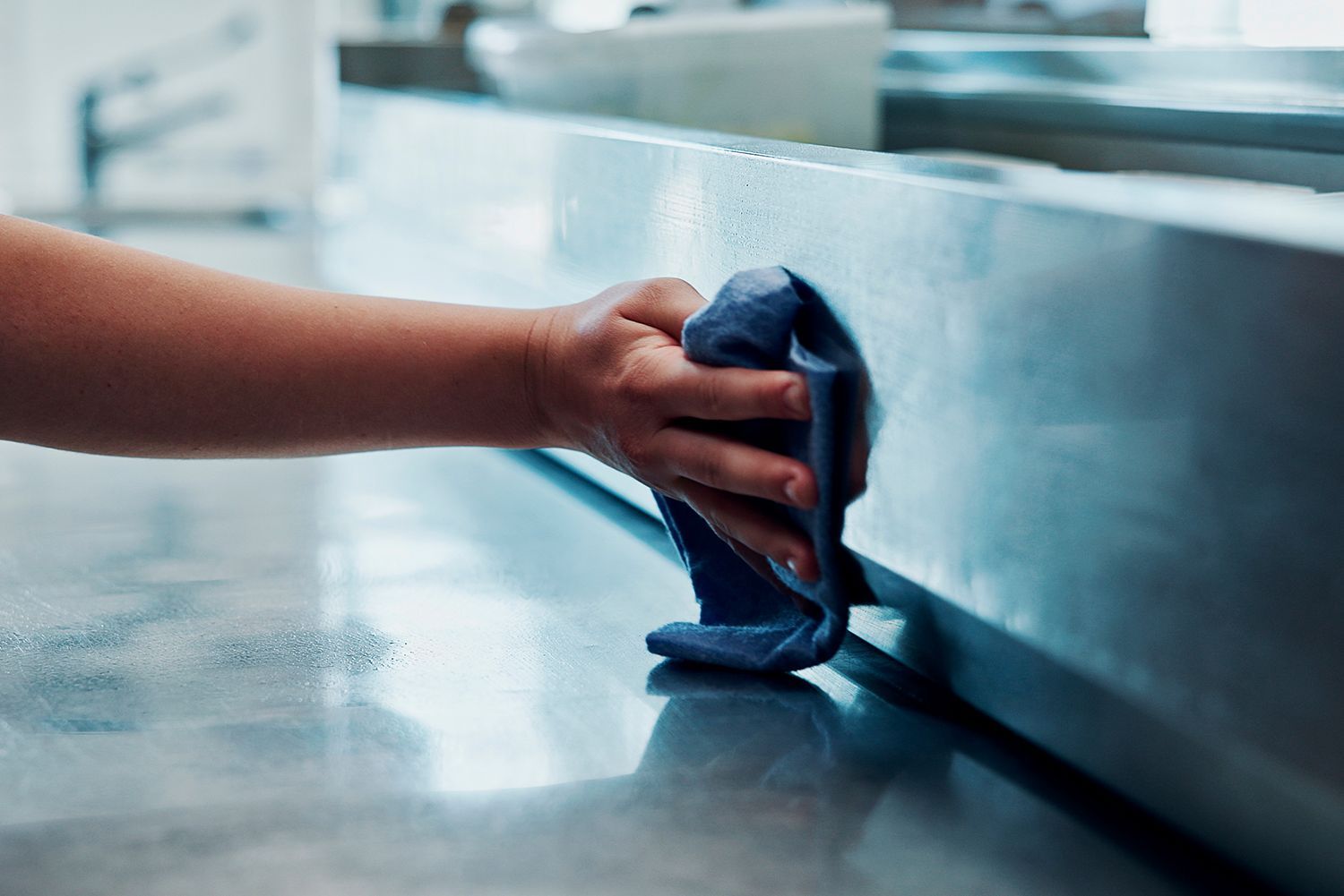 Hand wiping down a stainless steel countertop with a blue cloth.