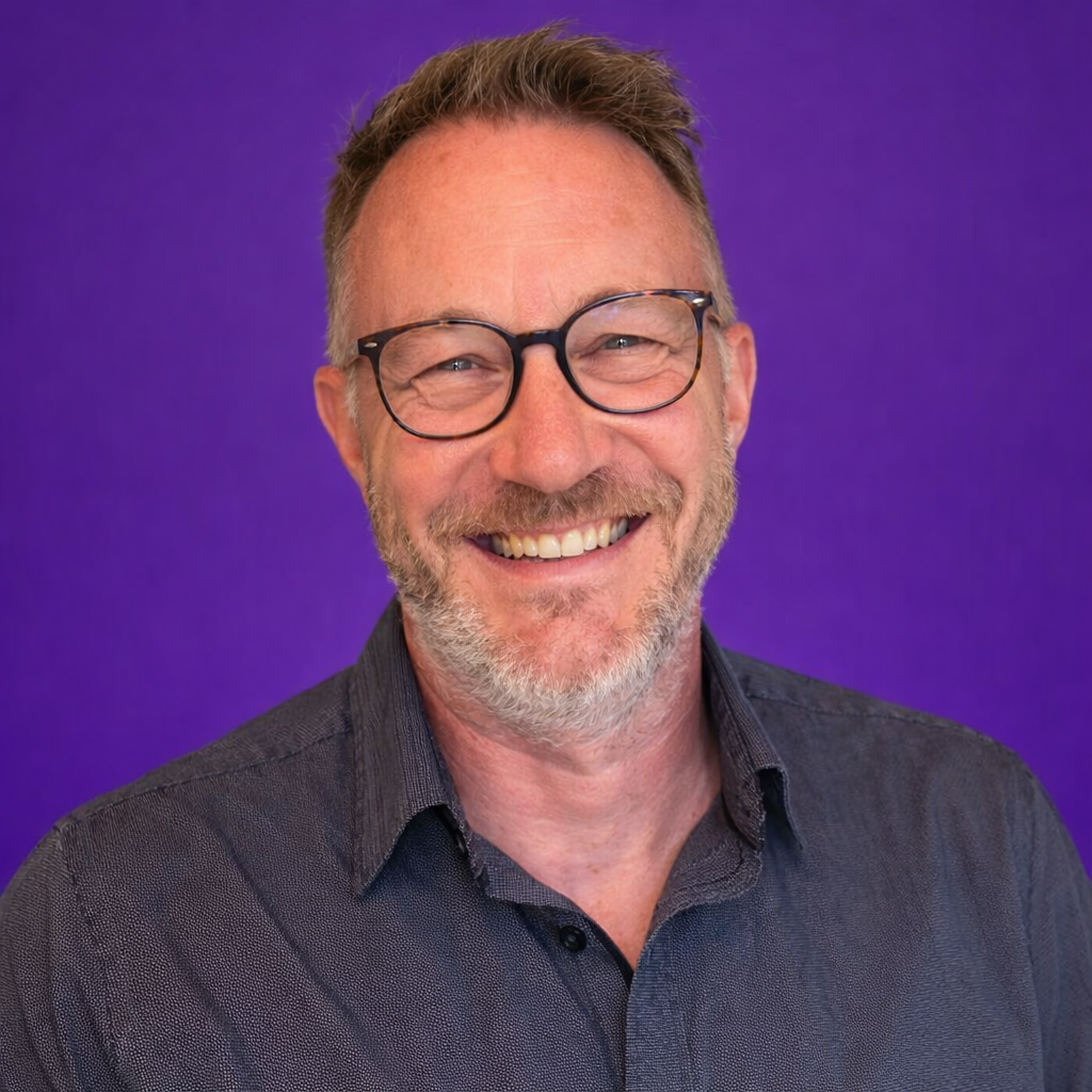 Man with glasses smiling, wearing a dark shirt, purple background.