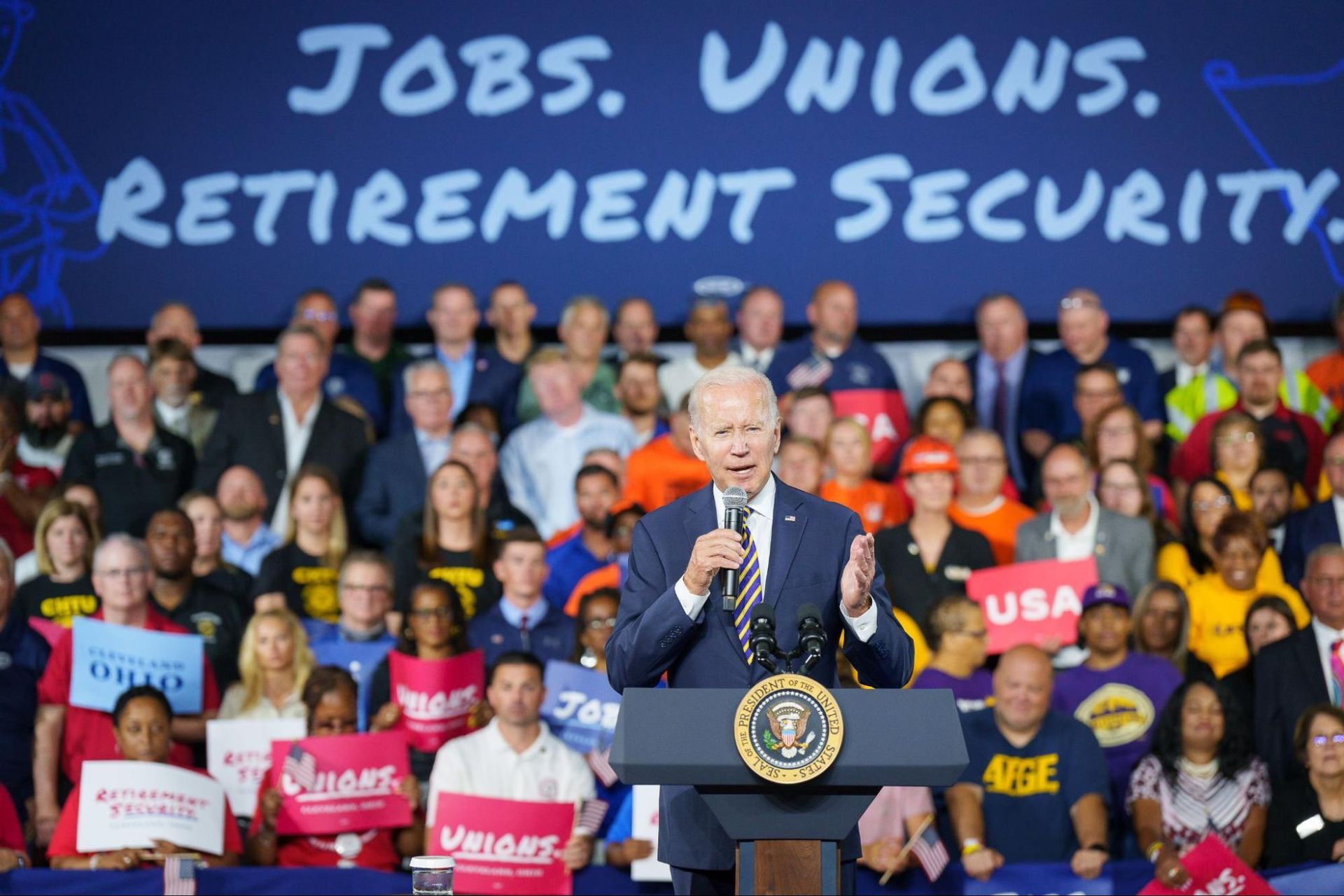 Biden speaking in front of a seated crowd with a banner above 