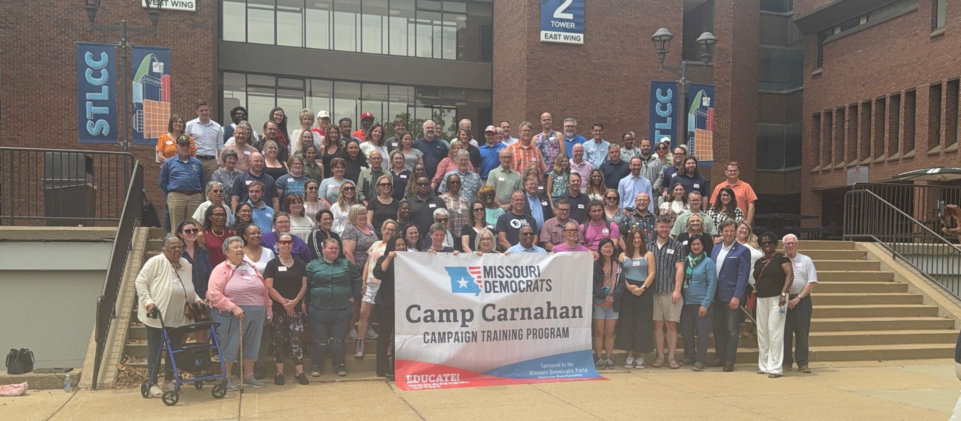 Group photo of over 100 attendees at Camp Carnahan, standing behind a banner on the steps of STLCC