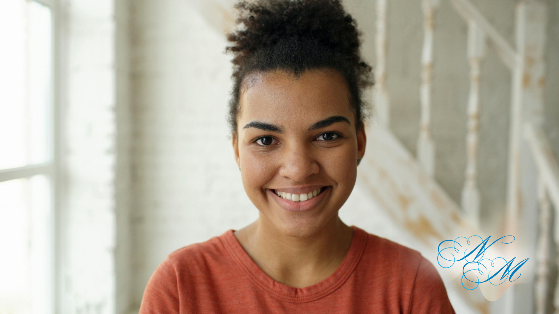 Smiling person in an orange shirt standing indoors with a bright, softly blurred background