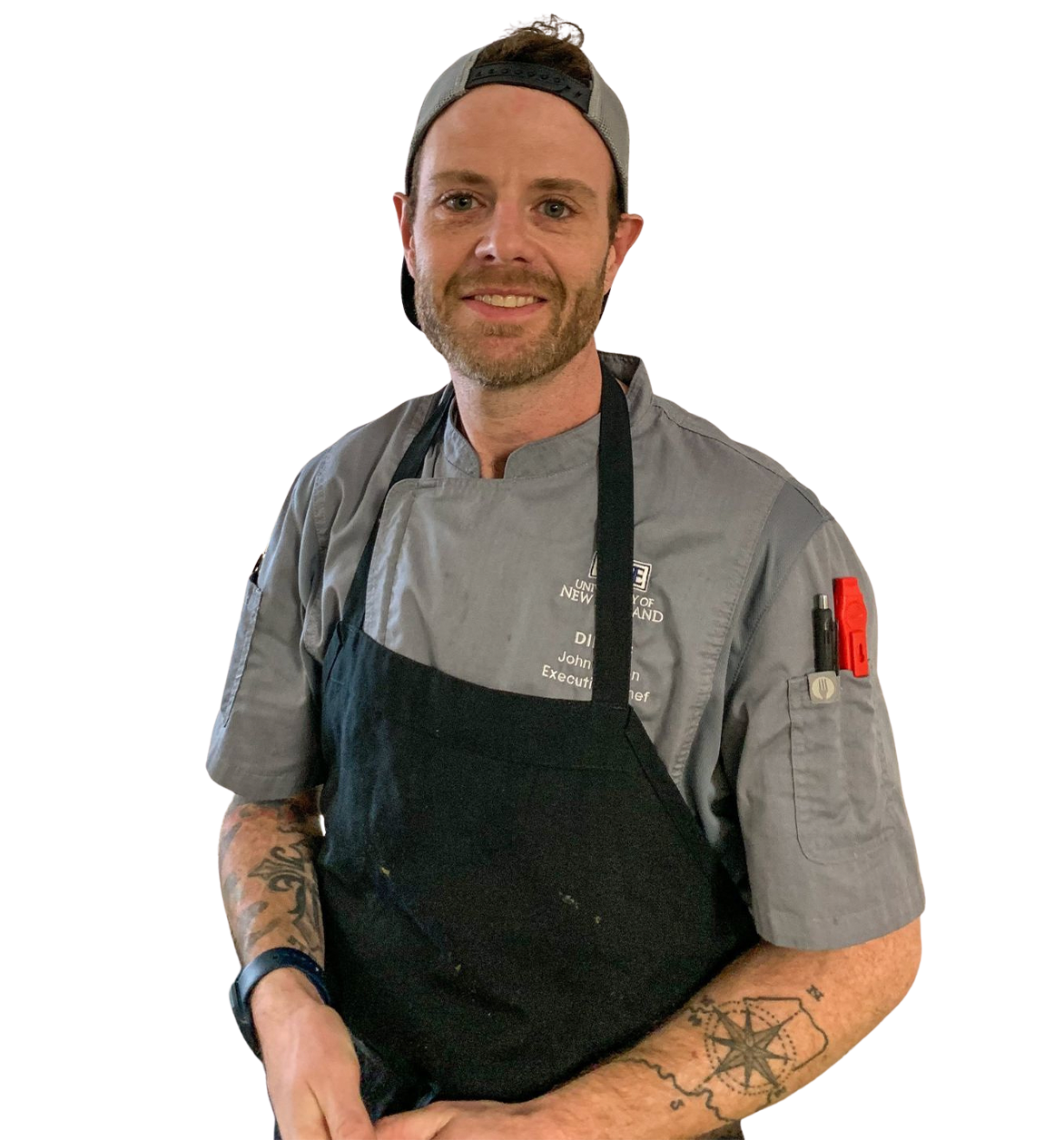 Chef smiling, wearing gray chef's coat and black apron, with tattoos and a baseball cap.