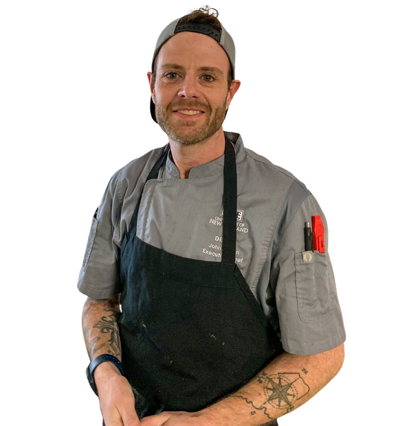 Chef smiling, wearing gray chef's coat and black apron, with tattoos and a baseball cap.