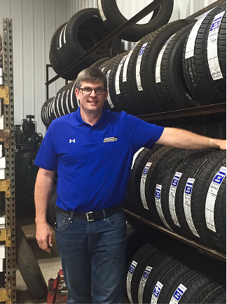 A person in a blue polo shirt stands in a warehouse aisle lined with shelves of black tires.