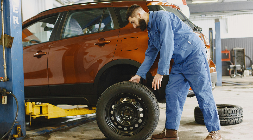 A mechanic in a blue jumpsuit maneuvers a car tire near a lifted orange SUV in an auto repair shop.