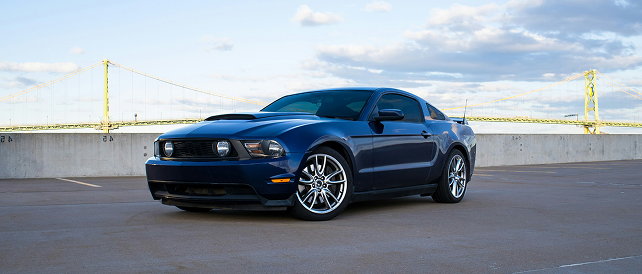 A dark blue Ford Mustang parked on an open-air rooftop with a suspension bridge visible in the background.