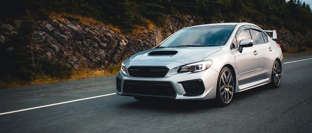 A silver Subaru WRX sports sedan driving on an asphalt road alongside a rocky hillside.