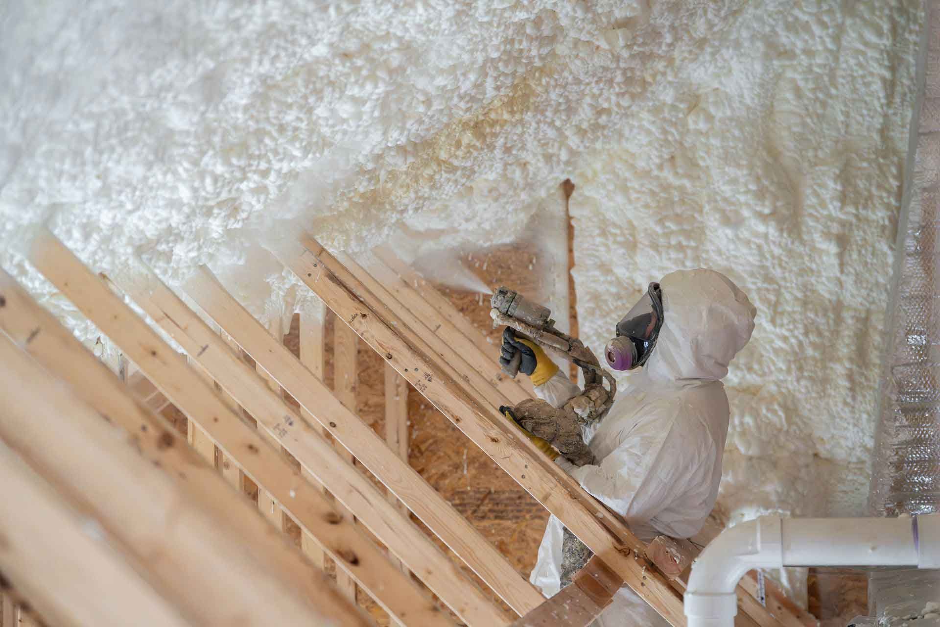 Worker in protective suit using spray foam insulation services to insulate attic wood and rafters.
