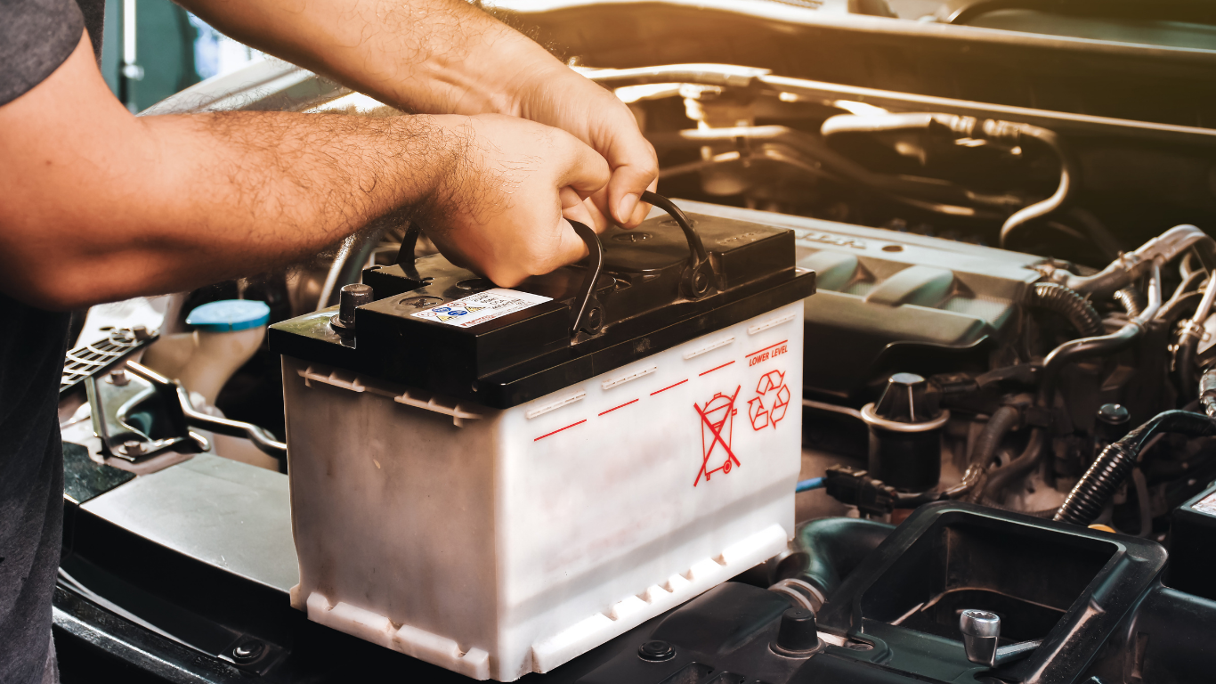 Person lifts a car battery from an engine, with a warning symbol on the battery.