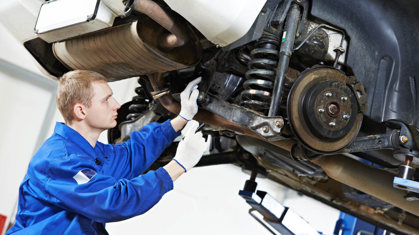 Mechanic in blue overalls working on the underside of a car in a garage.