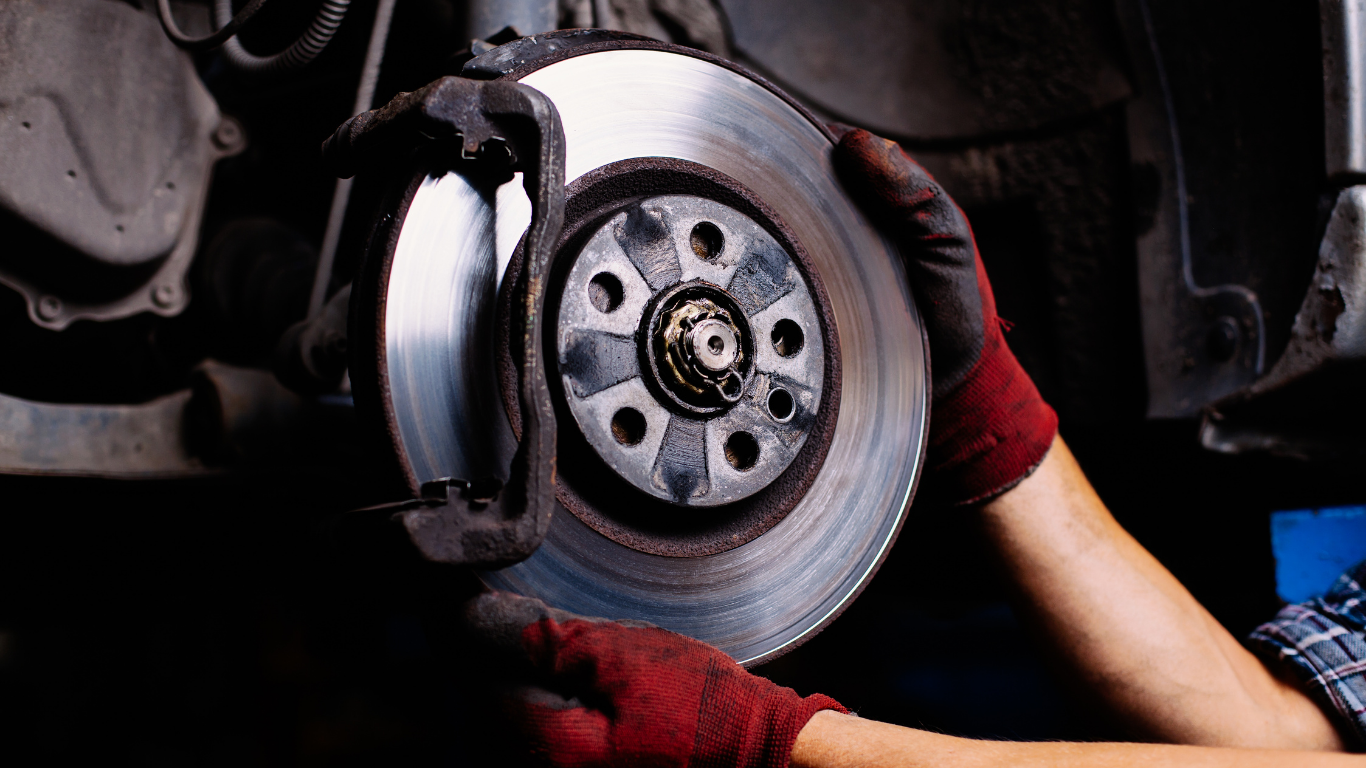 Hands in gloves holding a car brake rotor, in a mechanic's shop.