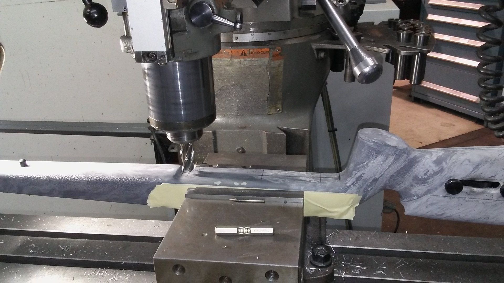 Machine tool drilling a metal bar in a workshop. Drill bit, vise, and tape visible.