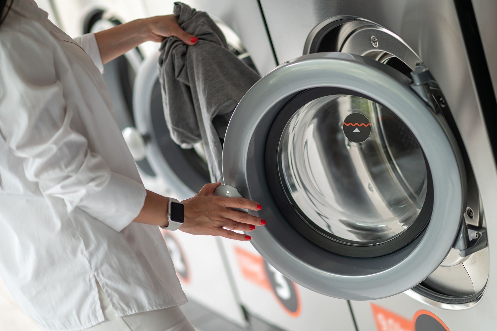Person loading a washing machine with gray clothing in a laundromat