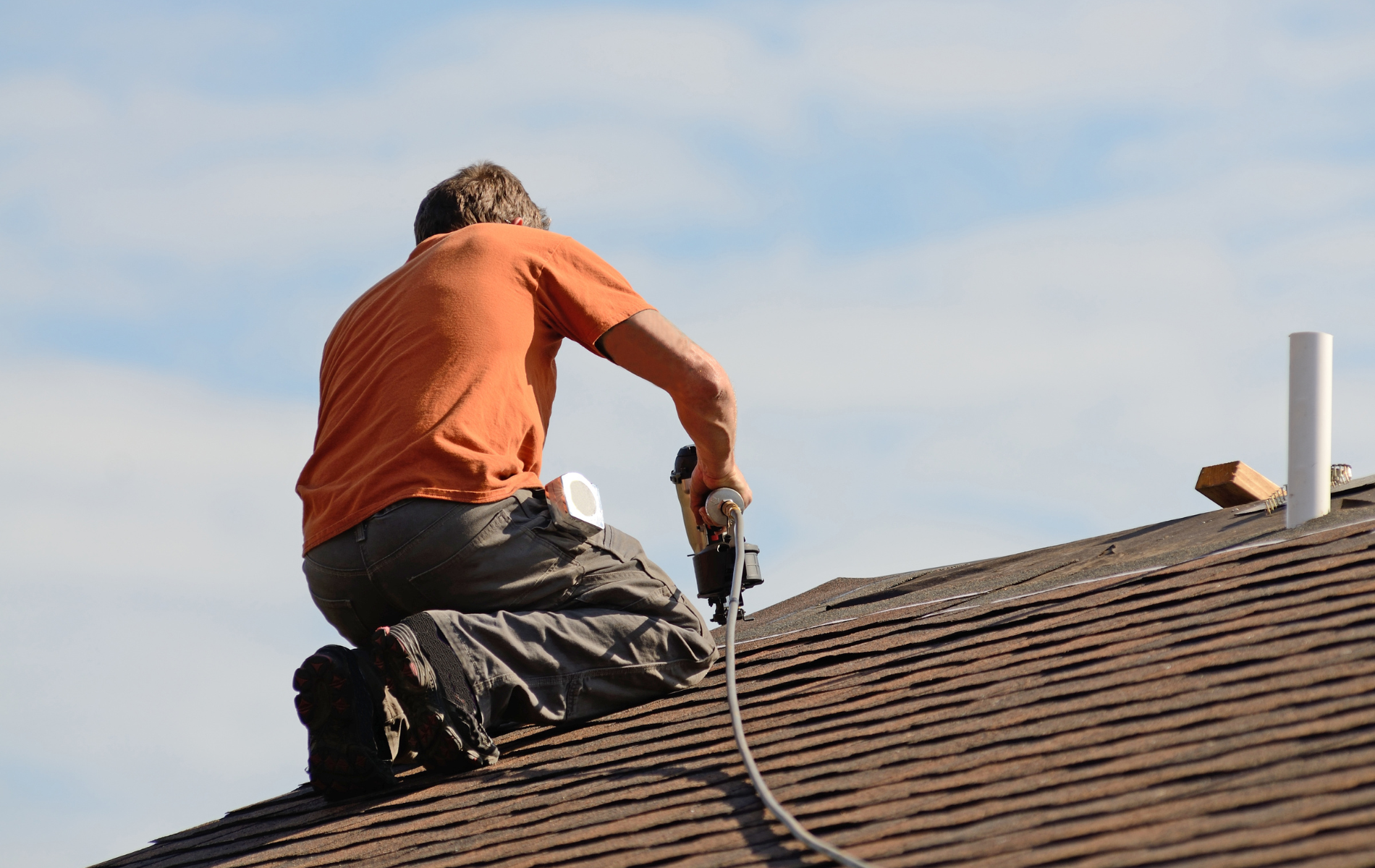 Roofer in orange shirt kneels on a brown shingle roof, using a nail gun under a blue sky.