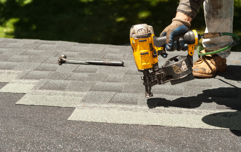 Person uses a nail gun to install roofing shingles on a rooftop. A hammer is nearby.