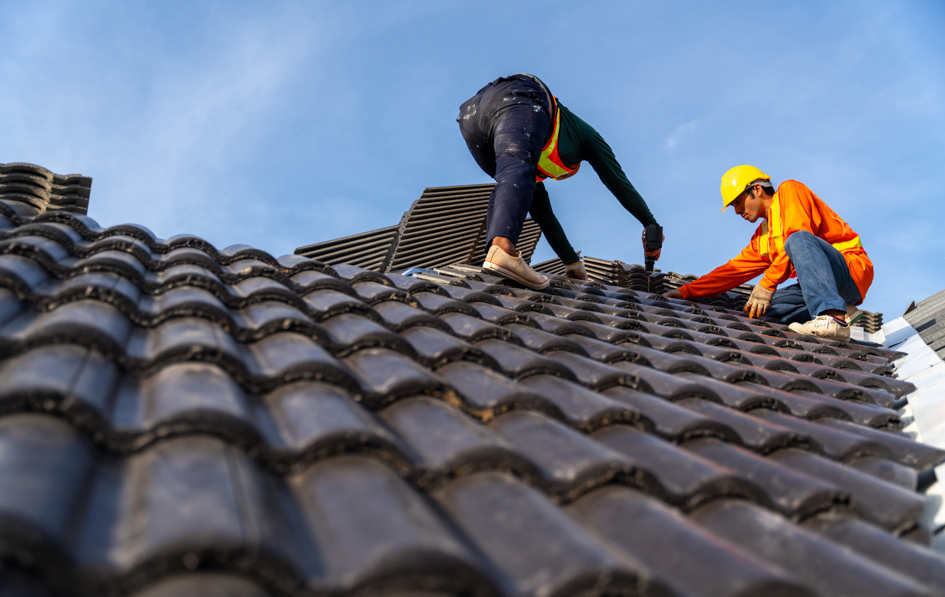 Two roofers installing dark gray tiles on a roof, blue sky backdrop.