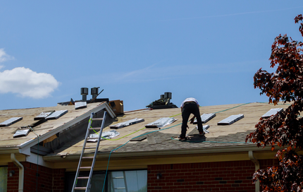 Person working on a roof, using a tool. Ladder nearby. Blue sky.