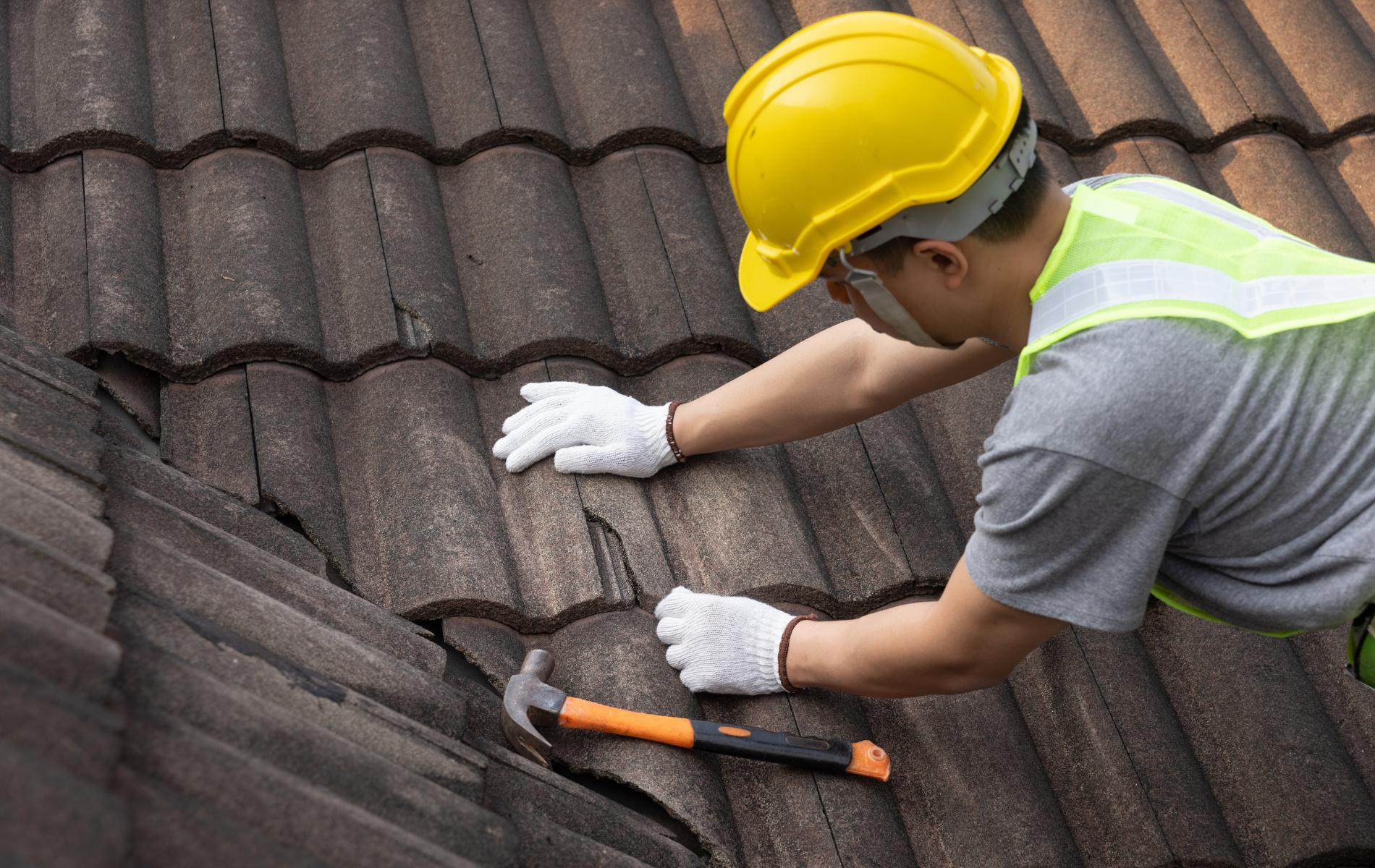 Roofer in yellow hard hat, safety vest, and gloves examines damaged roof tiles with a hammer.