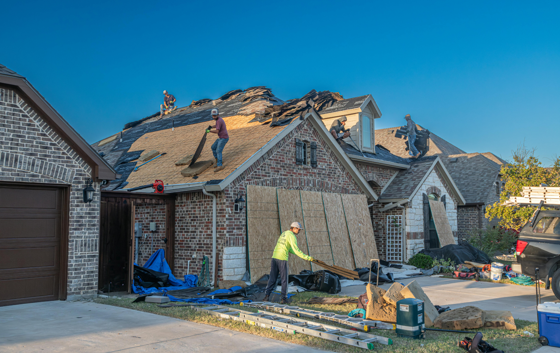 Roofers repairing a house with exposed plywood underlayment, various tools, and debris on a sunny day.