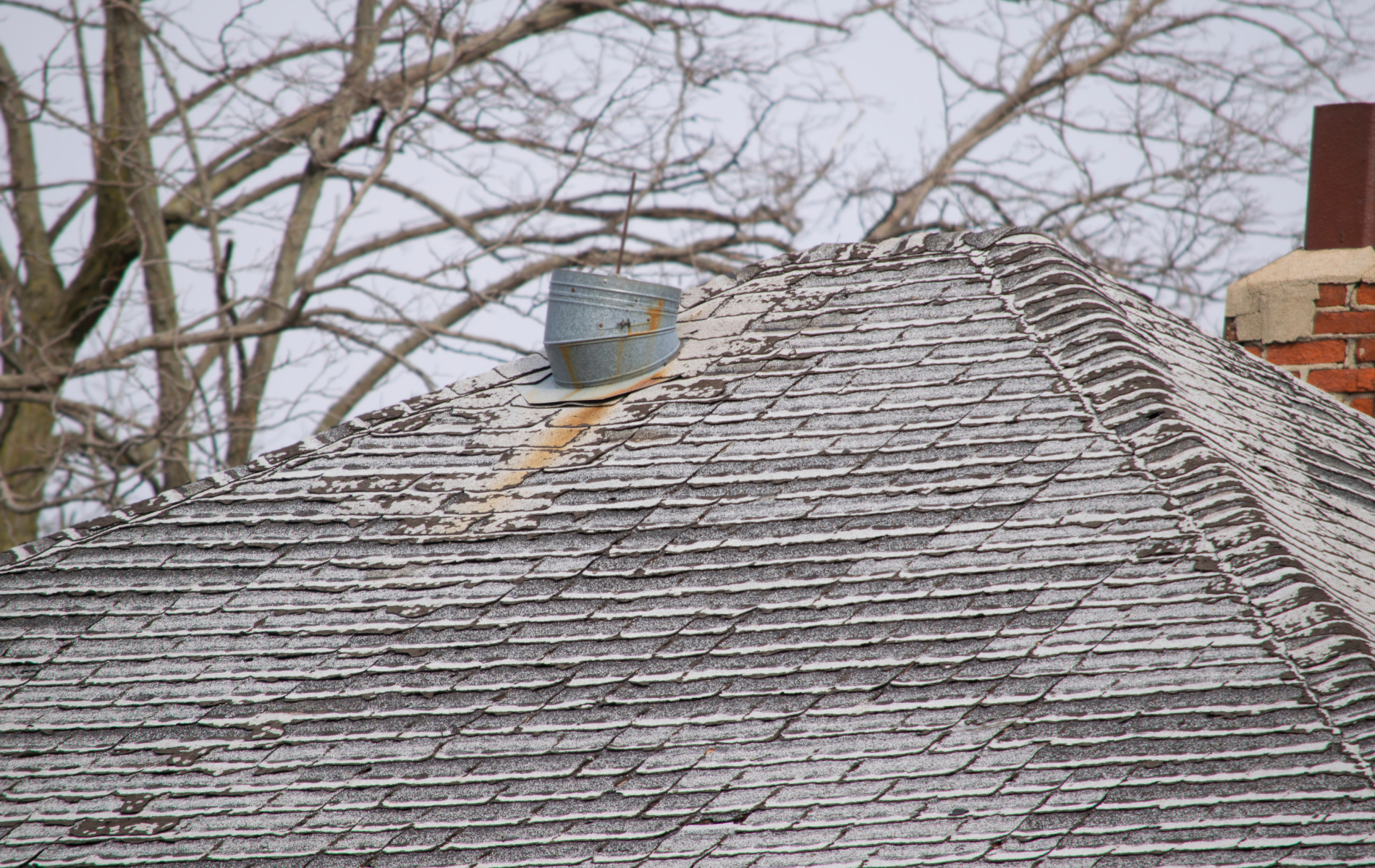 Old, weathered shingled roof with a vent and brick chimney in a winter setting with bare trees.