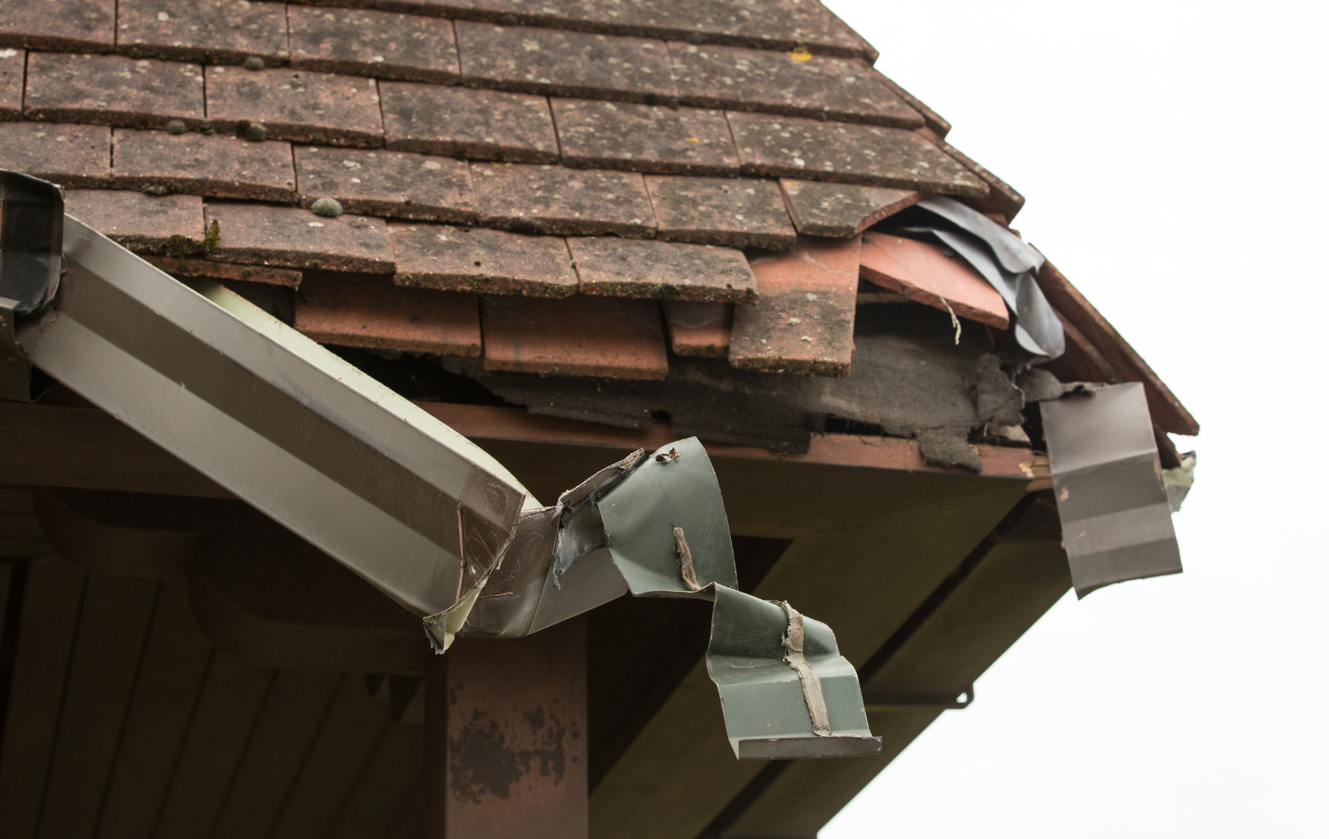 Damaged roof corner with broken tiles and crumpled guttering against a white sky.