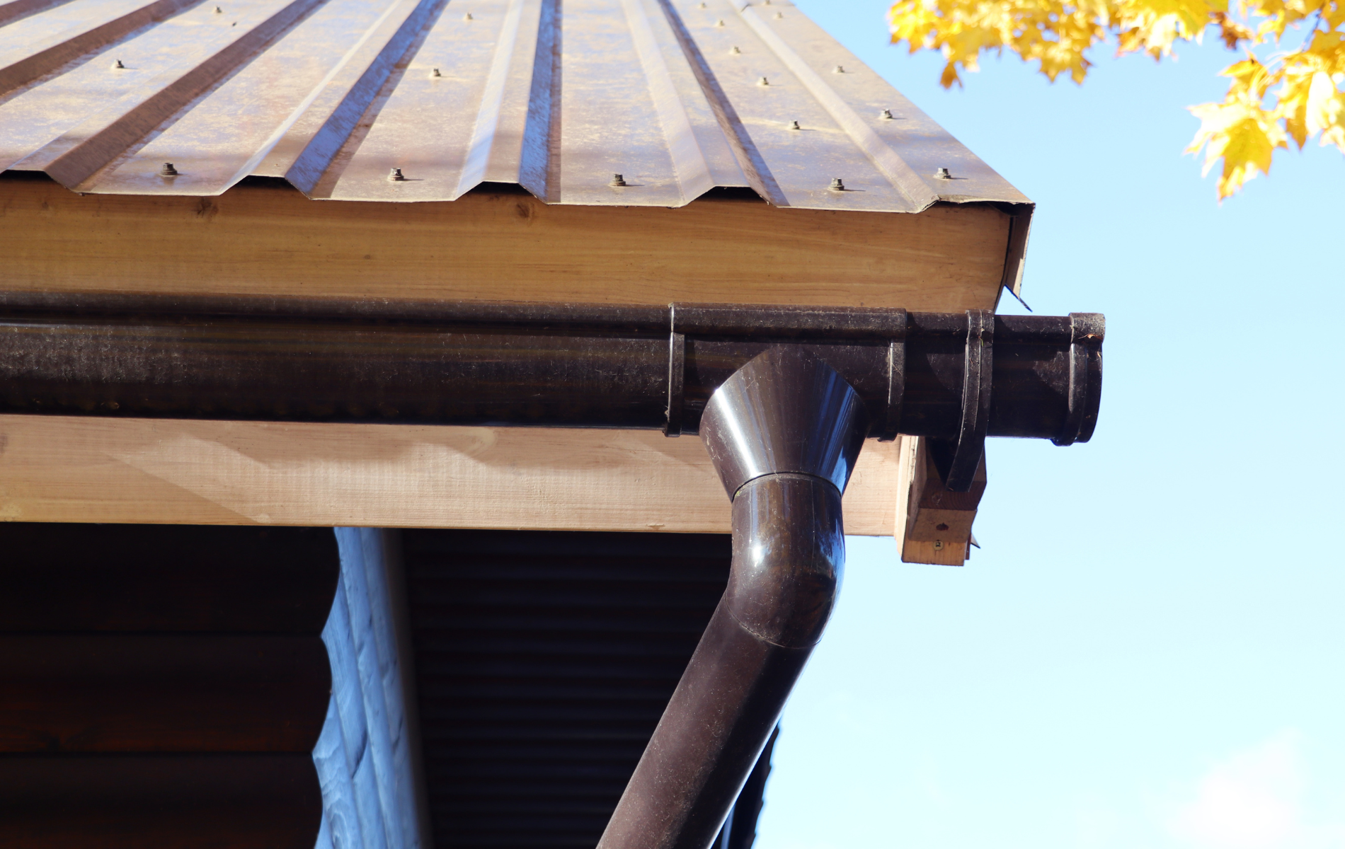 Brown rain gutter and downspout on a wooden roof with a metal roof panel, against a blue sky.