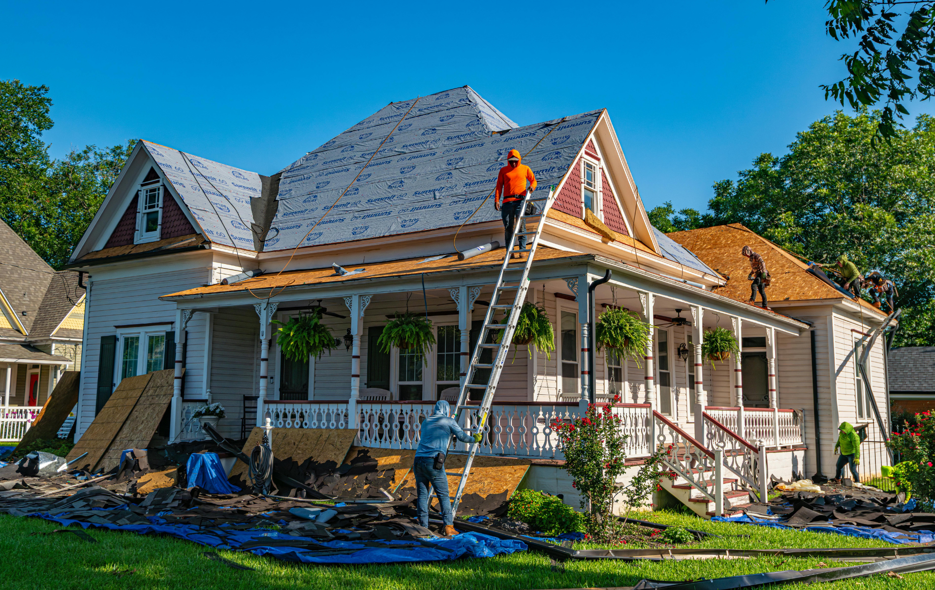 Roofers working on a two-story house with a porch; blue tarps and materials on the lawn.