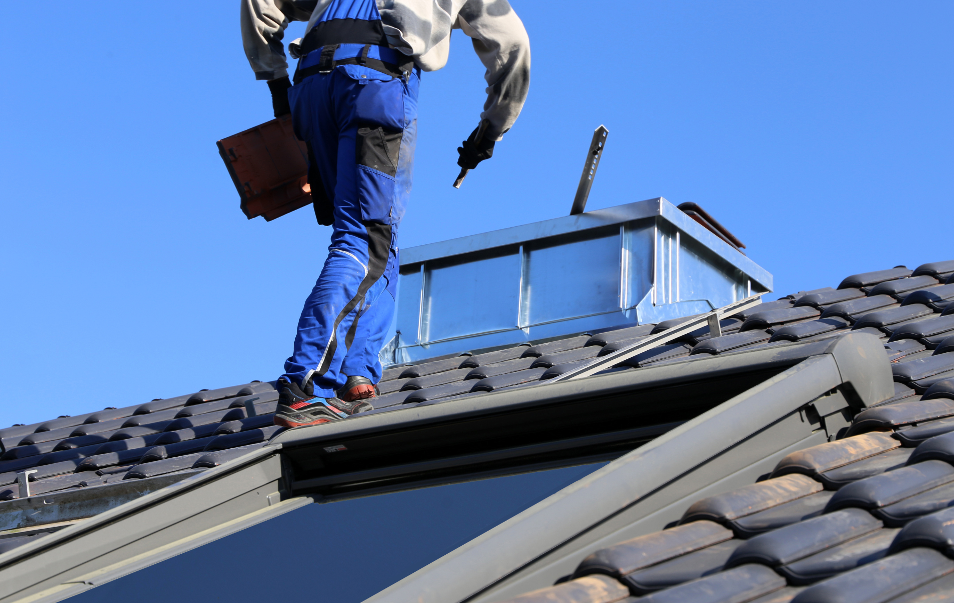 Roofer in blue overalls on a tiled roof working near a skylight on a sunny day.