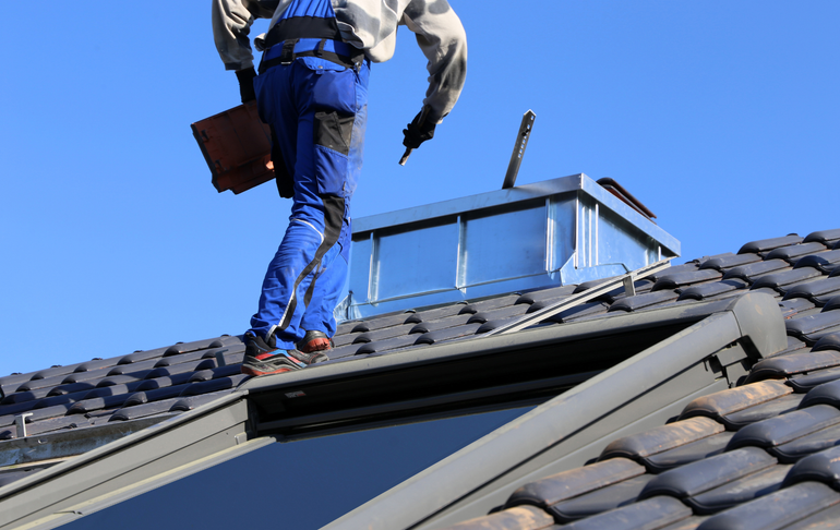 Roofer in blue overalls on a tiled roof working near a skylight on a sunny day.