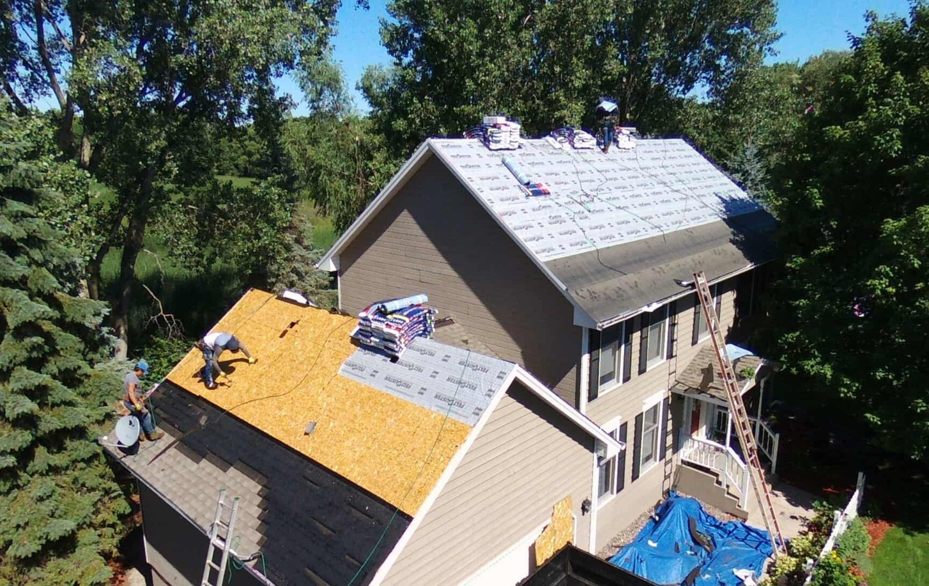 Roofers working on a two-story house, shingles partially installed. Blue tarp covers below.