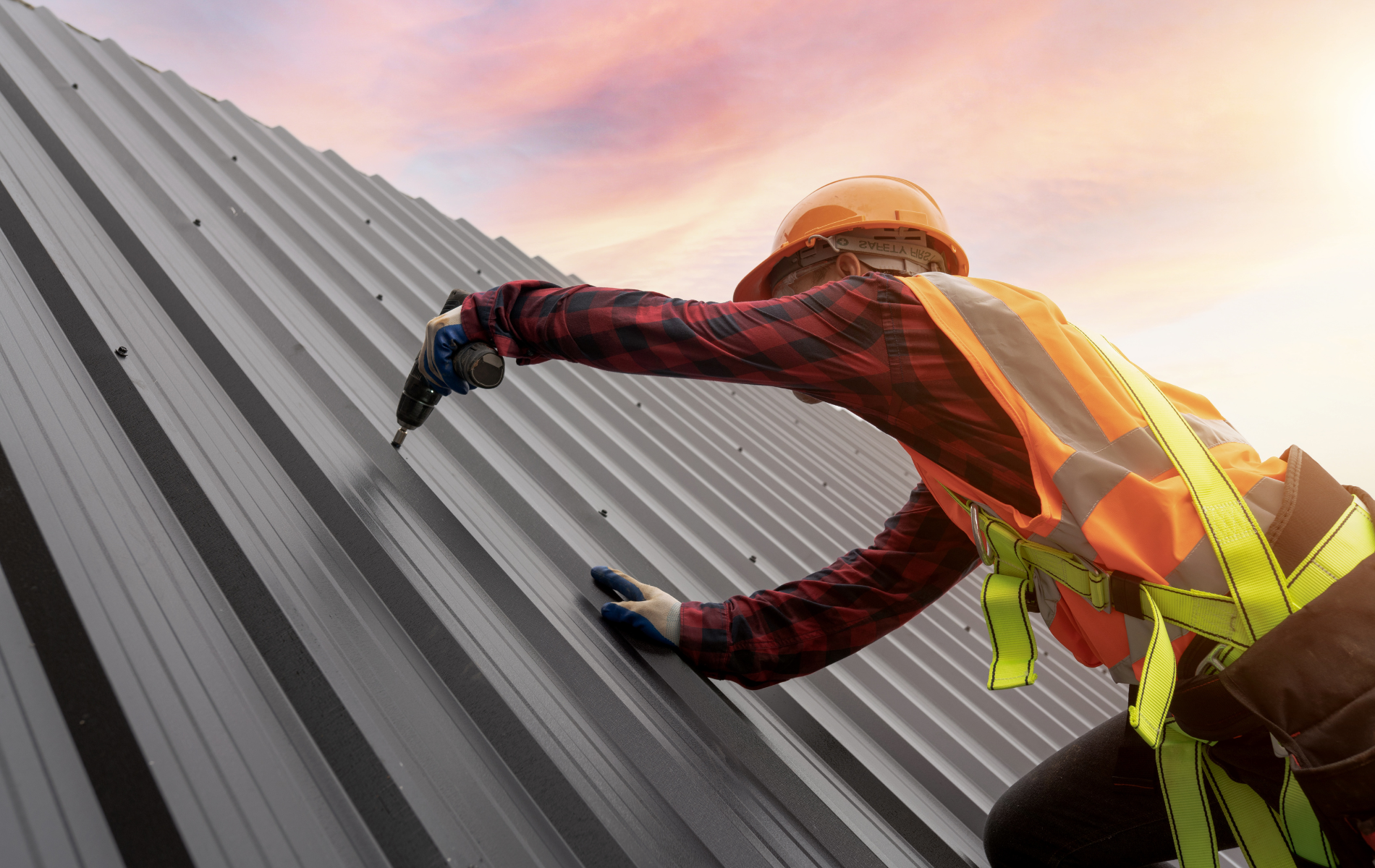 Roofer in safety gear installing metal panels on a sloped roof at sunset.