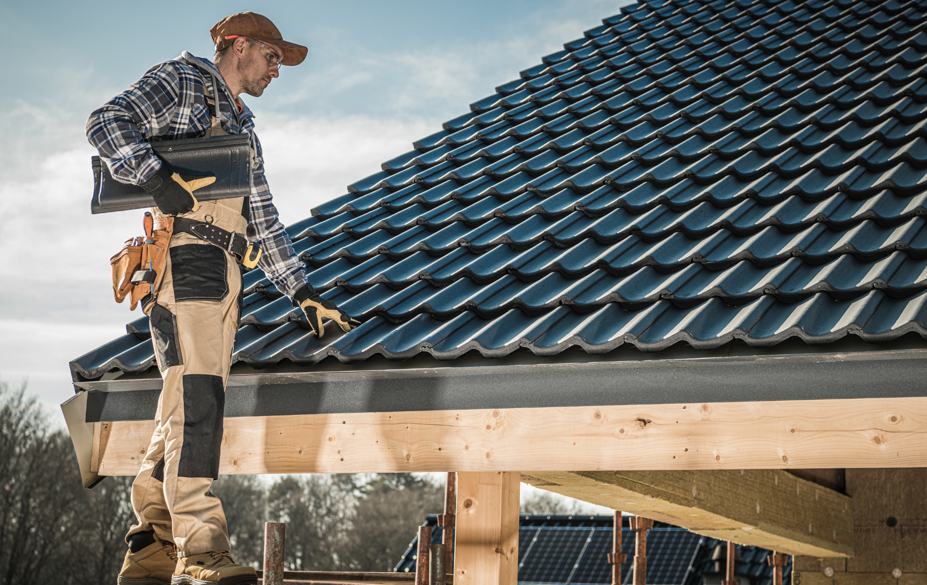 Roofer on a roof, inspecting dark tiles and a gutter. Man wears a hat, work clothes, and carries a tool.