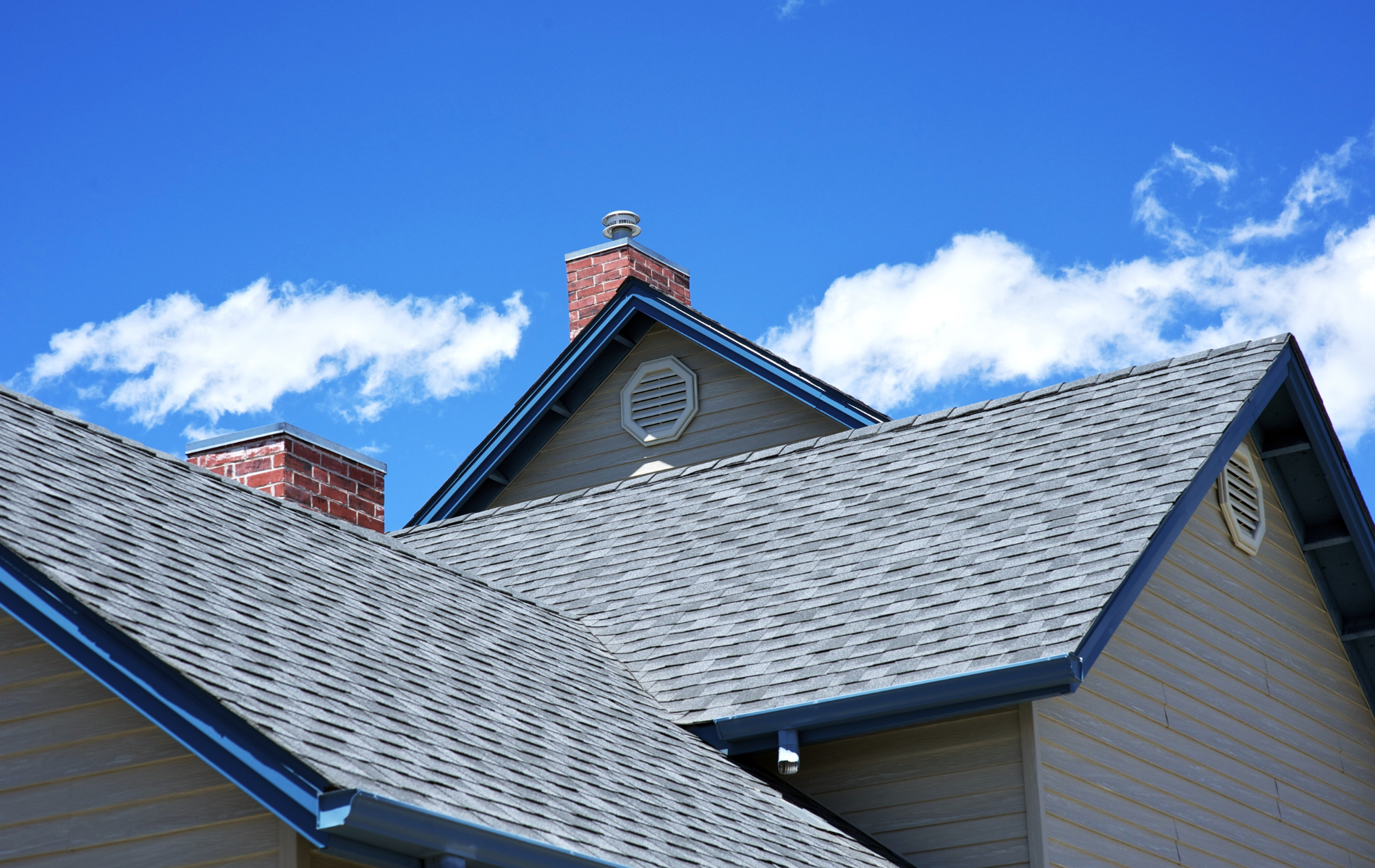 House roof with gray shingles, brown siding, and brick chimneys against a blue sky with clouds.