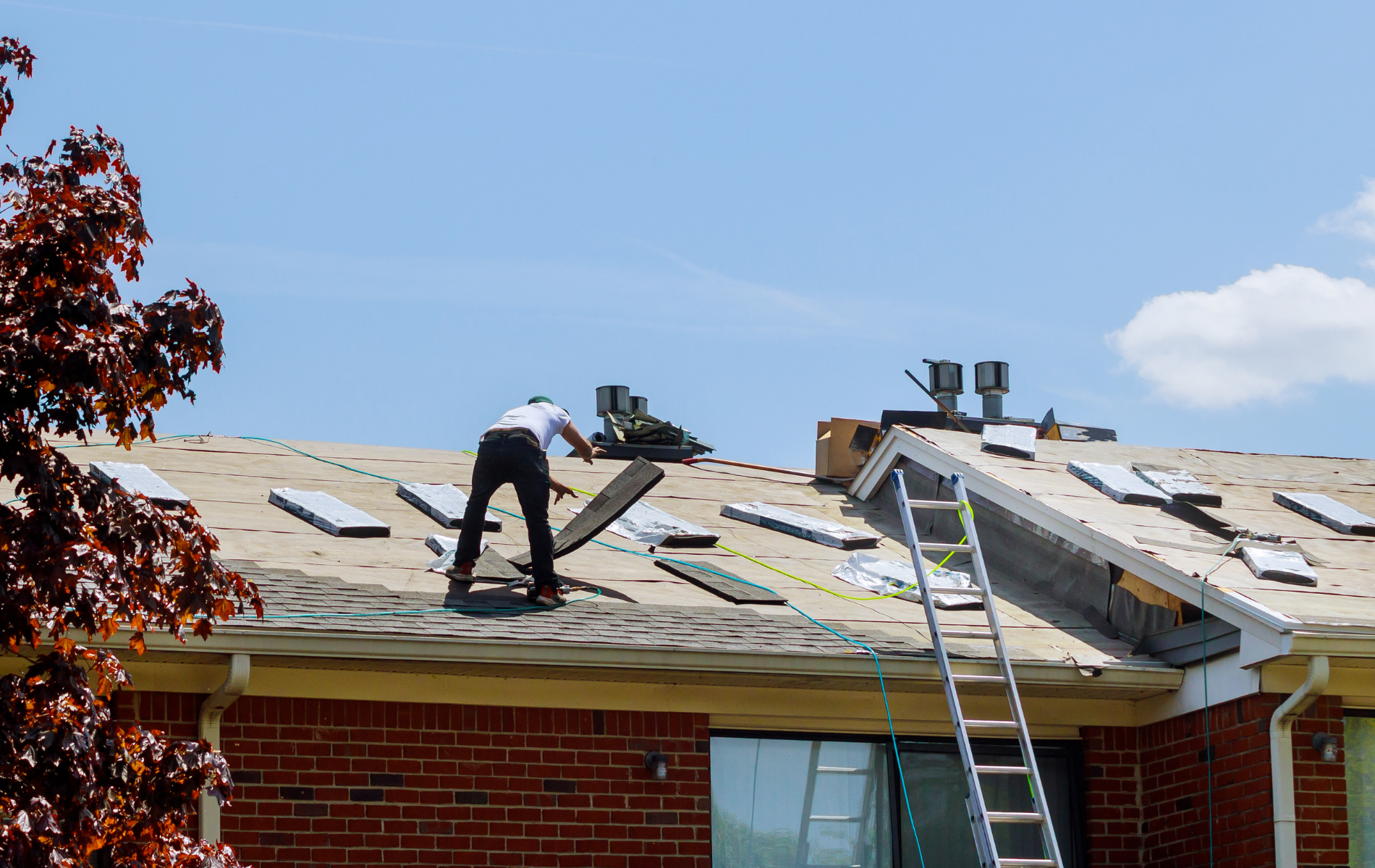 Roofer on a roof, replacing shingles. A ladder is propped against the roof. Blue sky in the background.