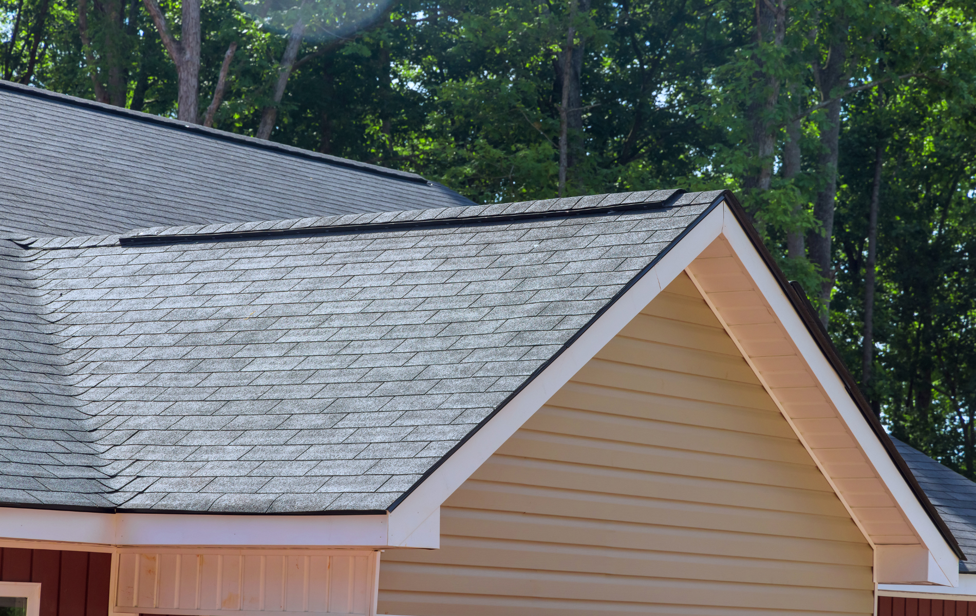 Roof of a house with gray shingles and beige siding against a backdrop of green trees.